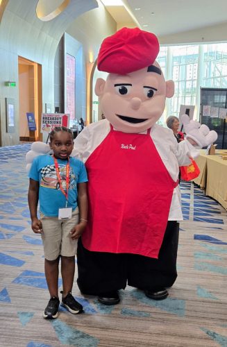 A young African American attendee in a blue shirt standing with The Family Cafe mascot.
